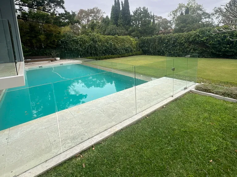 Pool behind a frameless glass fence, with a lawn and tall hedges around the yard
