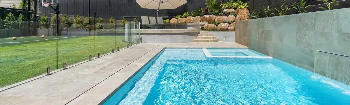 Backyard pool behind a frameless glass pool fence, with a lawn, palm trees and a dark timber boundary fence