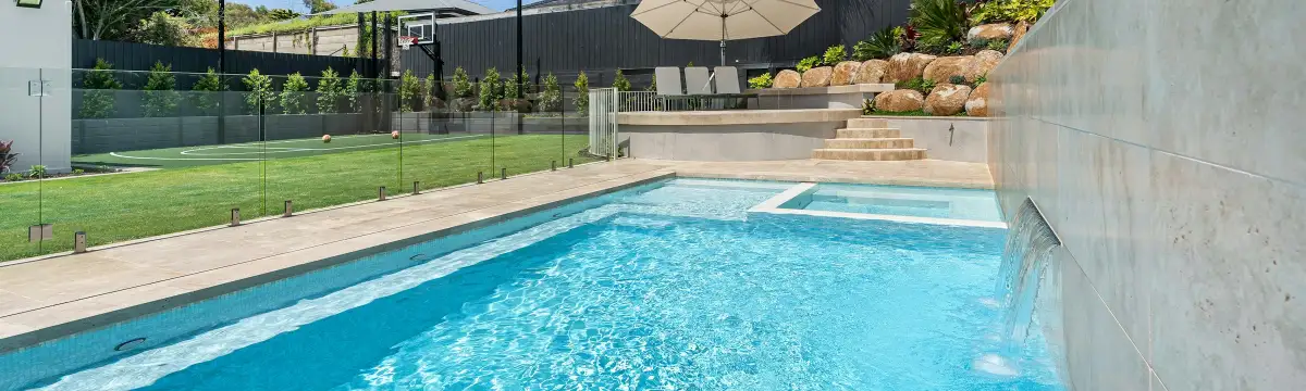 Rectangular backyard pool with pale stone coping and a frameless glass barrier along the lawn